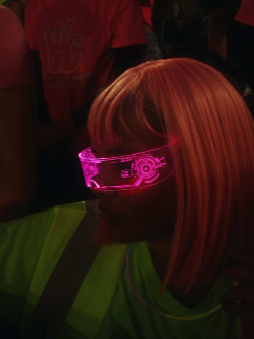A teenager wearing a bright pink neon wig, wraparound sunglasses, and a neon safety vest during a school spirit day.