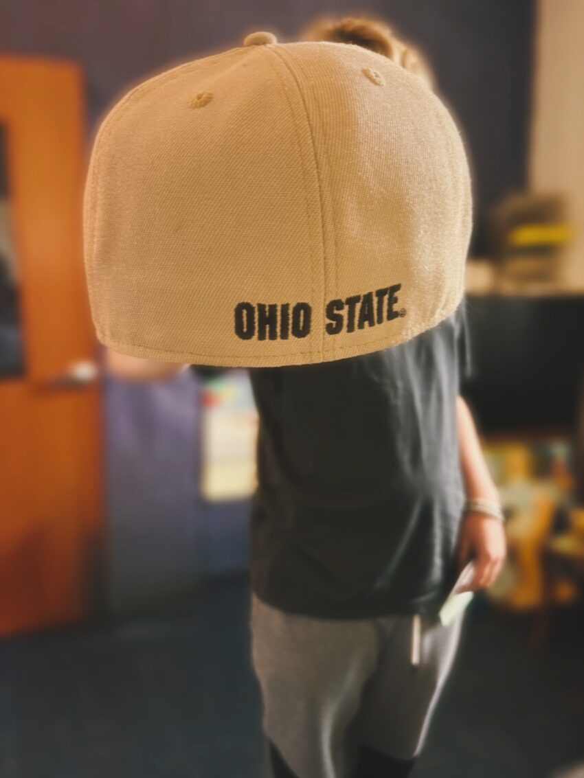 A person holding an Ohio State baseball cap forward, with the back of the hat visible while the face is intentionally out of frame.