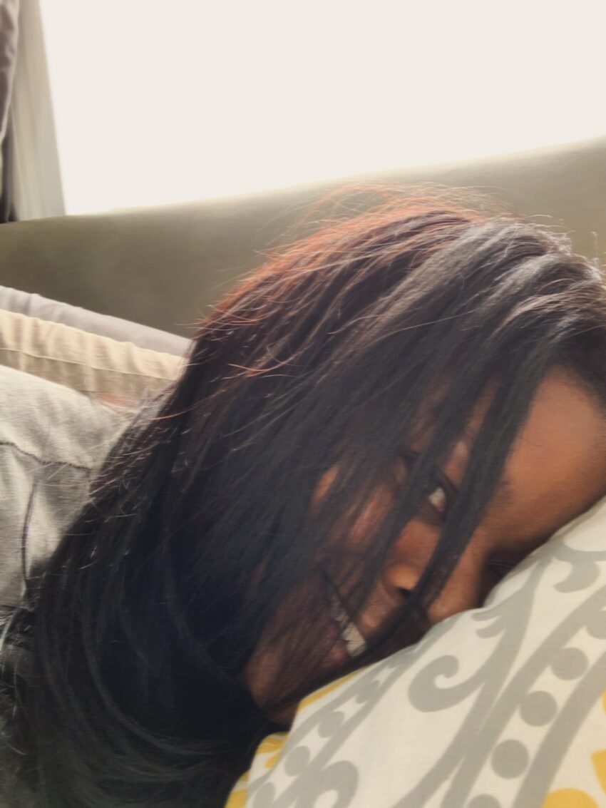 Close-up of a woman lying on a bed with long hair partially covering her face, one eye visible, and smiling playfully at the camera.