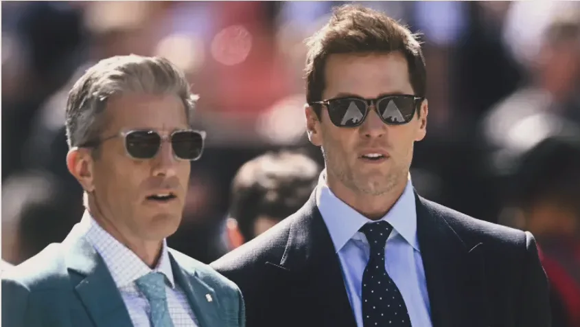 Tom Brady and Kevin Burkhardt standing together on the field before an NFL game.