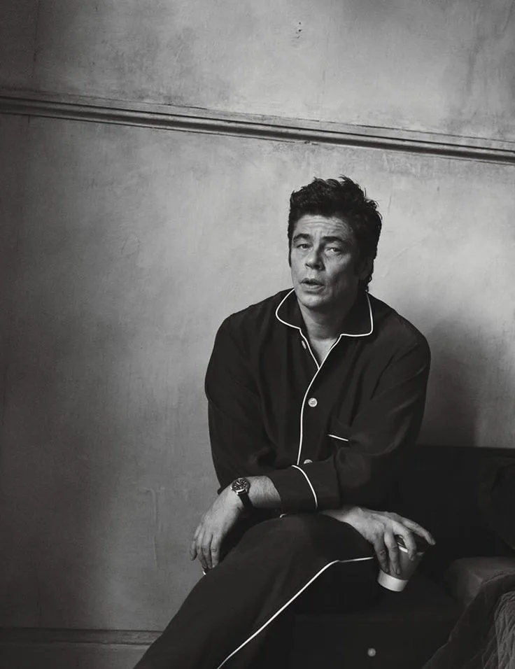 Black-and-white portrait of a Benicio del Toro wearing dark pajamas with white piping, seated and holding a paper cup of coffee.