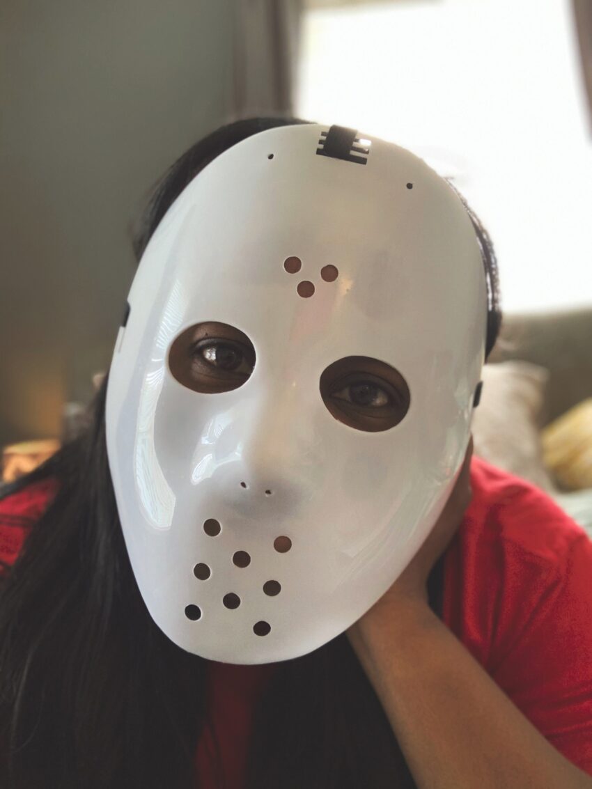 Woman in red shirt wearing Jason Voorhees mask, lounging on bed.