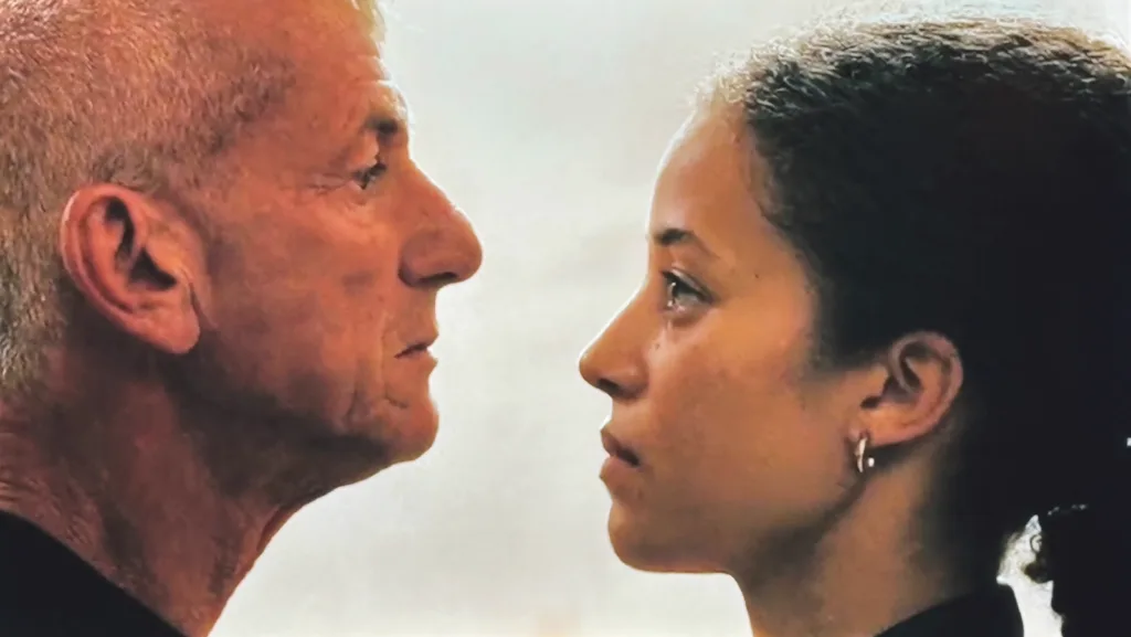 A young woman stands face to face with a man, holding his gaze in a tense, quiet moment.