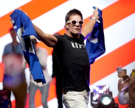 Tom Brady at Fanatics Fest wearing sunglasses and holding up a destroyed Eli Manning jersey, smiling and celebrating in a playful moment against a vibrant orange and blue background